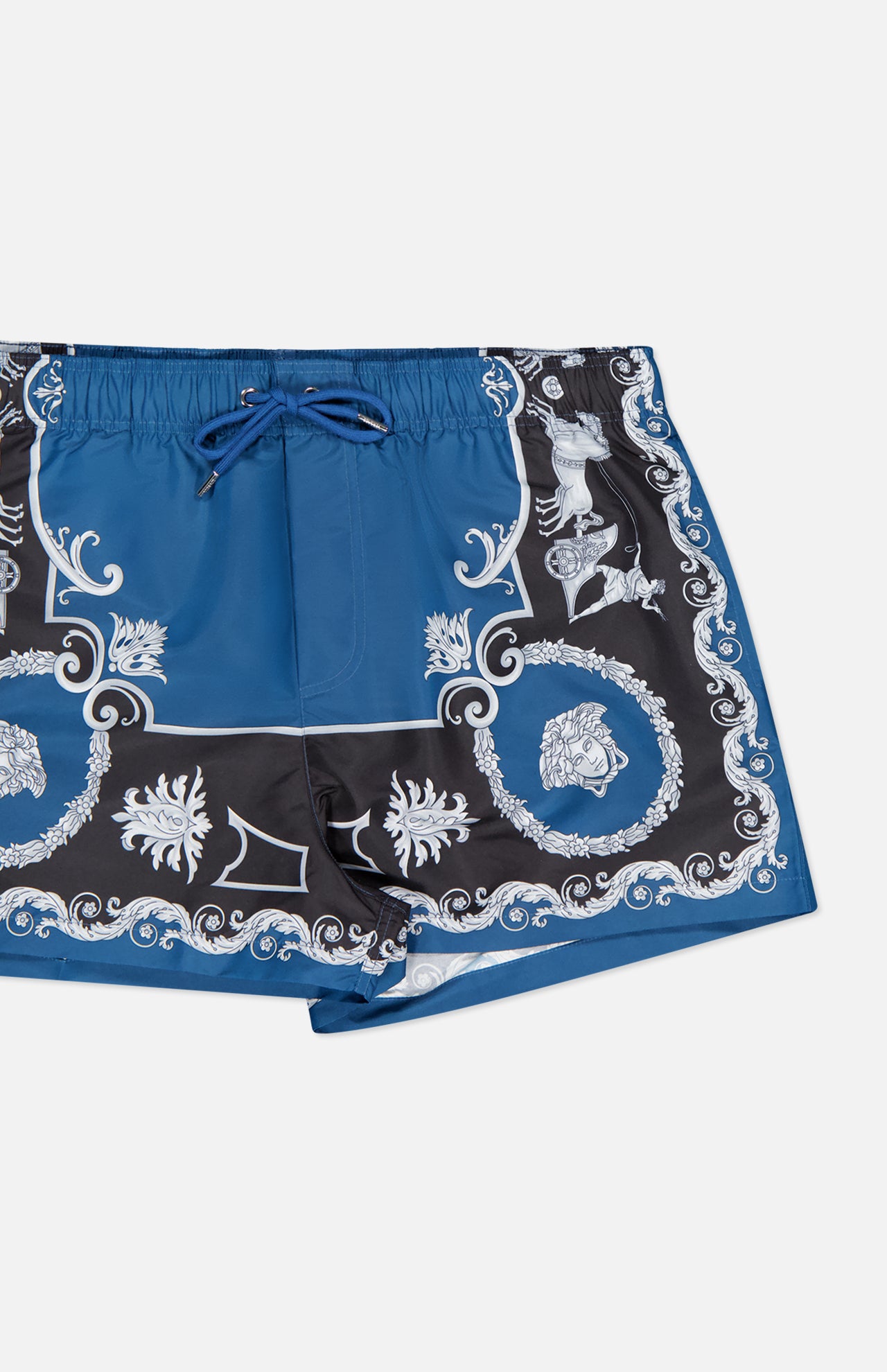 The VERSACE Mythic Barocco Print Swim Shorts feature a blue and black baroque design with white ornate floral and scroll motifs, an elastic waistband, and a blue drawstring. Shown here against a white background.