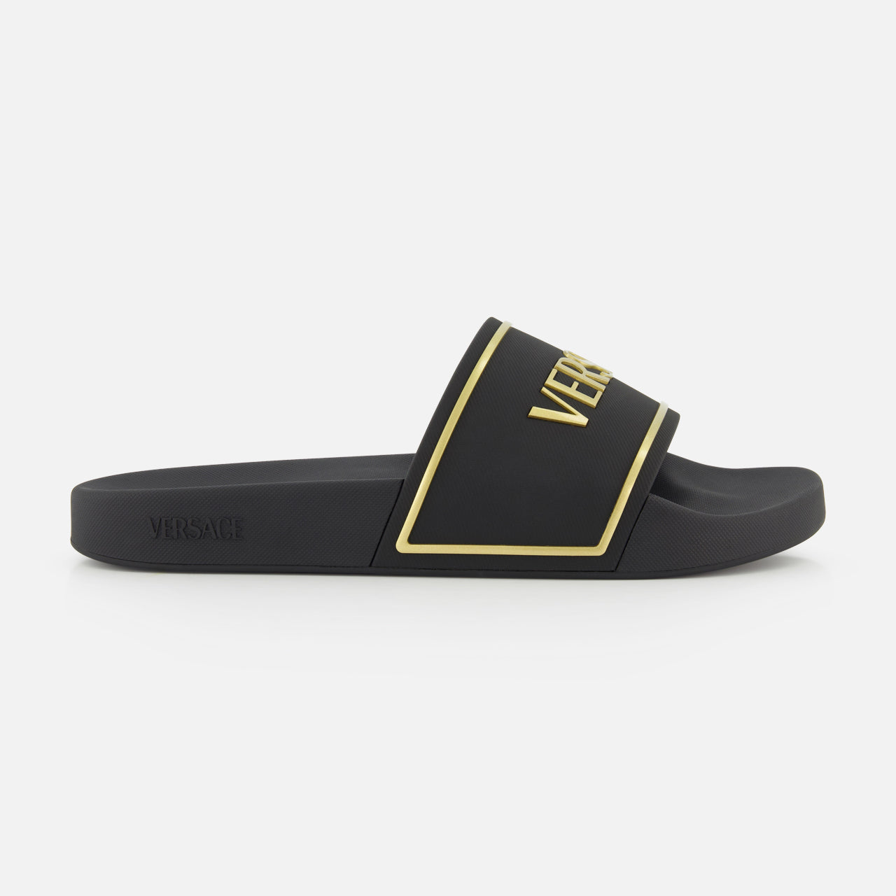 The Rubber + Embossing Versace Slide Sandals by VERSACE feature a wide black strap with gold trim and bold branding, minimalist design, and flat sole, elegantly displayed from the side against a white background.
