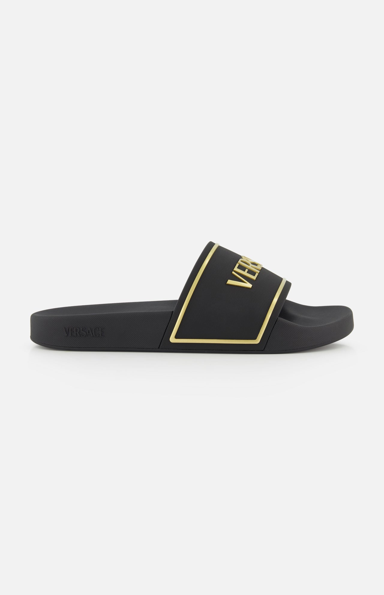 The Rubber + Embossing Versace Slide Sandals by VERSACE feature a wide black strap with gold trim and bold branding, minimalist design, and flat sole, elegantly displayed from the side against a white background.