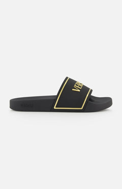 The Rubber + Embossing Versace Slide Sandals by VERSACE feature a wide black strap with gold trim and bold branding, minimalist design, and flat sole, elegantly displayed from the side against a white background.