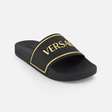 The Rubber + Embossing Versace Slide Sandals by VERSACE feature a wide black strap with gold trim and bold VERSACE lettering, showcasing luxury Italian style against a plain white background.