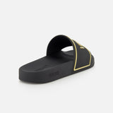 The VERSACE Rubber + Embossing Slide Sandal in black and gold features a wide embossed strap with gold details and logo, shown on a white background at an angle highlighting the side and interior—luxury Italian footwear.