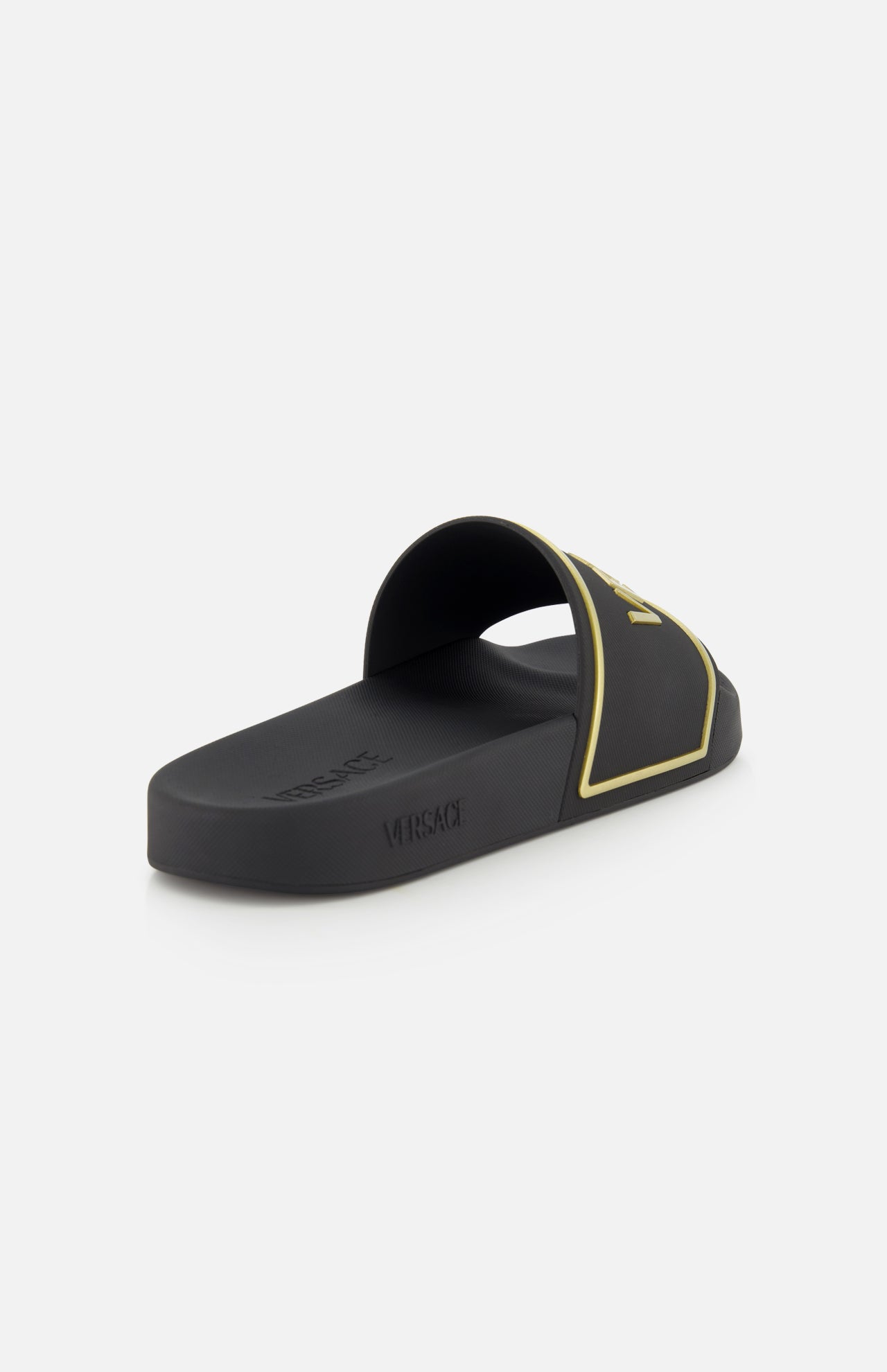 The VERSACE Rubber + Embossing Slide Sandal in black and gold features a wide embossed strap with gold details and logo, shown on a white background at an angle highlighting the side and interior—luxury Italian footwear.