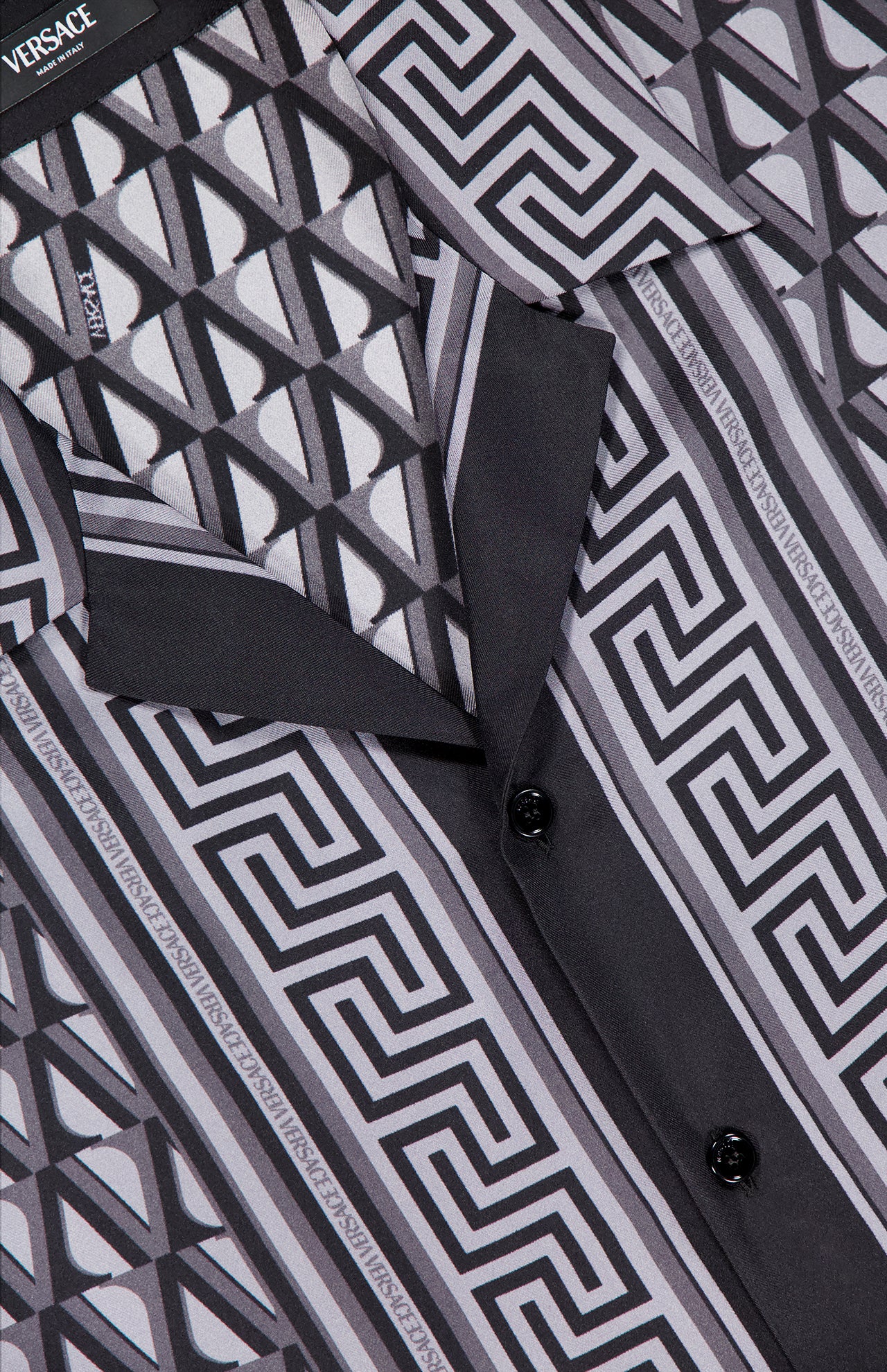 Close-up of the VERSACE V Greca Print Silk Shirt, a slim-fit men's shirt featuring a bold black, white, and gray geometric pattern with interlocking lines, black buttons, and a visible collar.