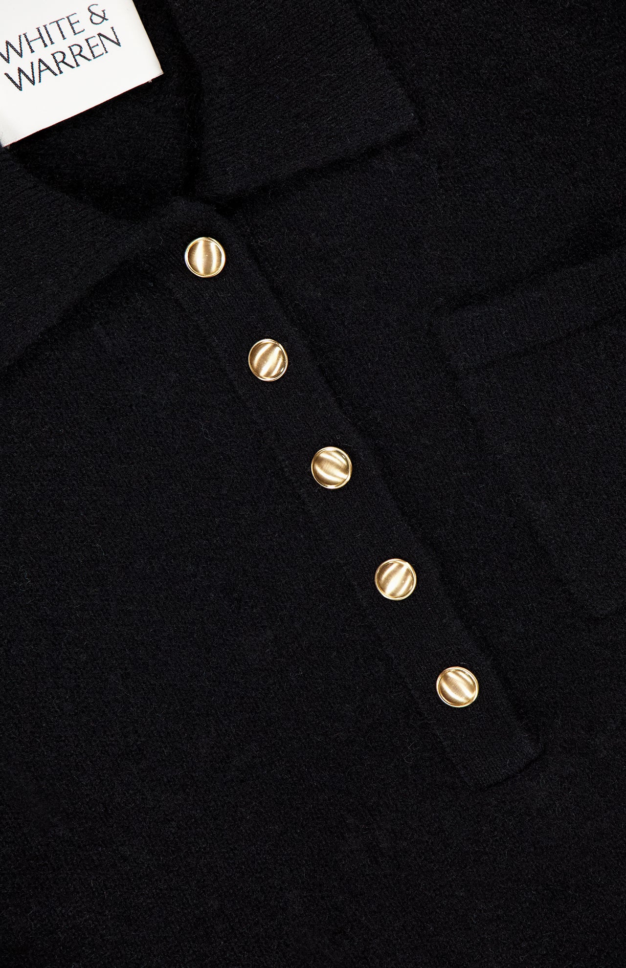 Close-up of the WHITE + WARREN Cashmere Gold Button Polo Top in black, featuring four gold-tone buttons and a small chest pocket; “WHITE + WARREN” label visible in the top left corner.