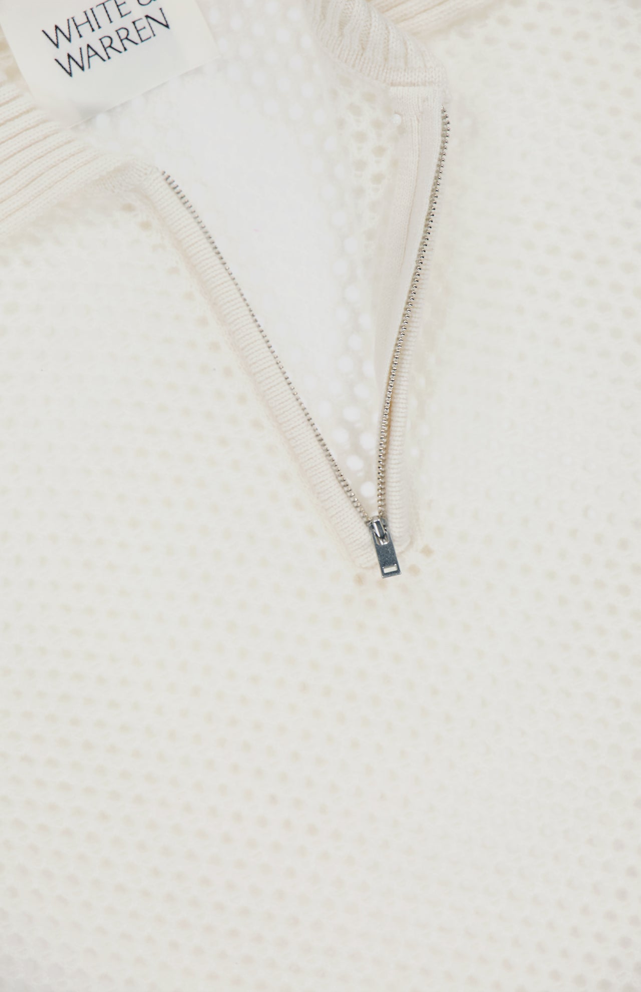 Close-up of the WHITE + WARREN Cashmere Mesh Polo Shirt, featuring a textured off-white mesh construction. The partially unzipped neckline reveals inner fabric, while a tag in the top left corner displays the brand name.
