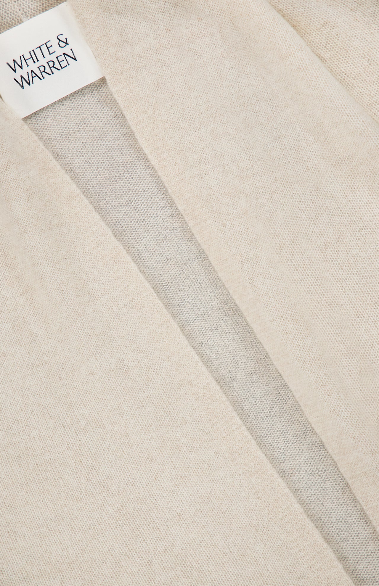 Close-up of a soft beige knit fabric with a visible seam and a WHITE + WARREN label sewn into the neckline, highlighting the luxurious texture of the Cashmere Mini Trapeze Cardigan in a slim fit.