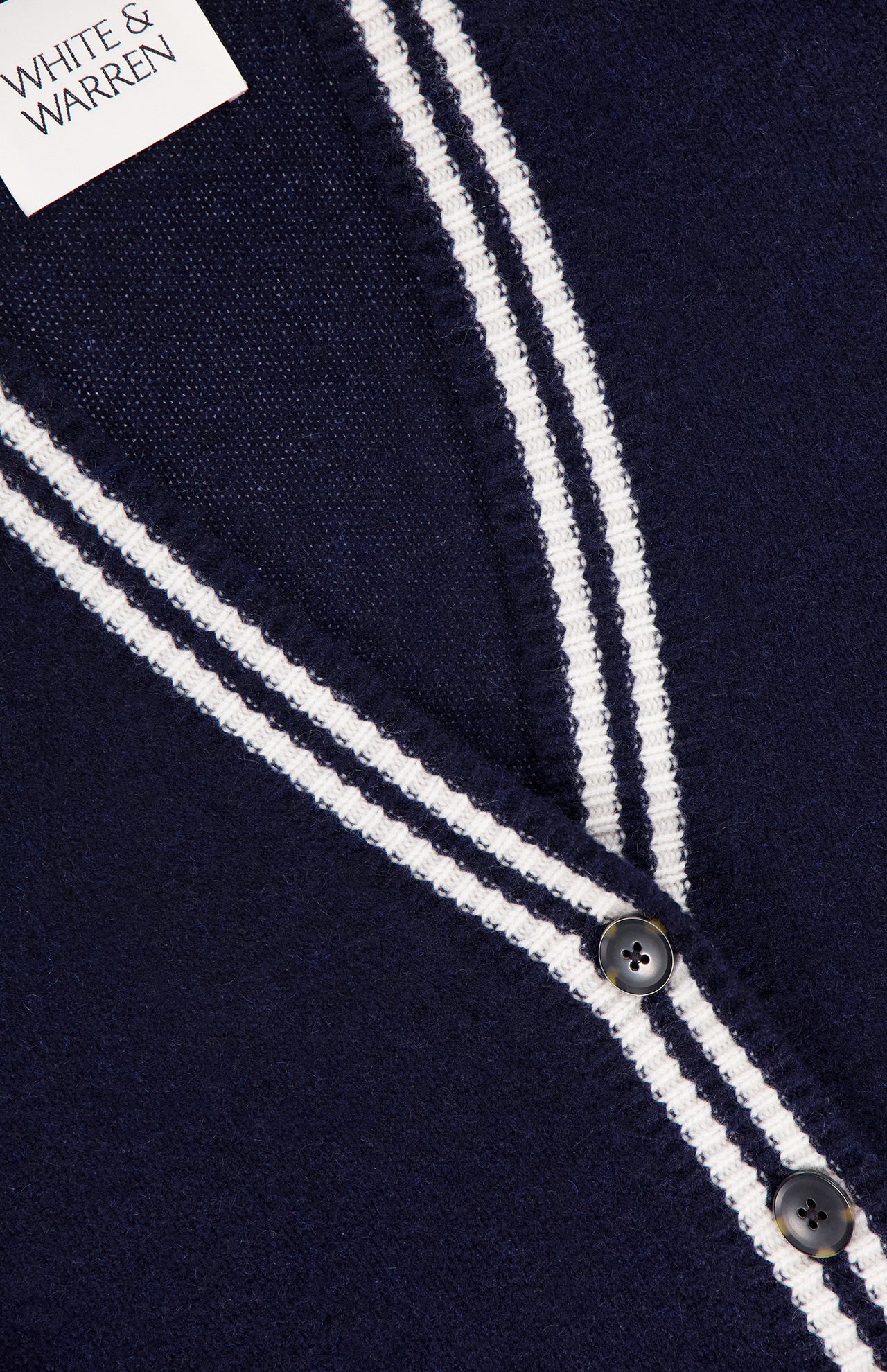 Close-up of the WHITE + WARREN Cashmere Varsity Cardigan in navy, featuring white double stripes on the V-neckline and button placket, dark buttons, and a visible WHITE + WARREN label in the top left corner.