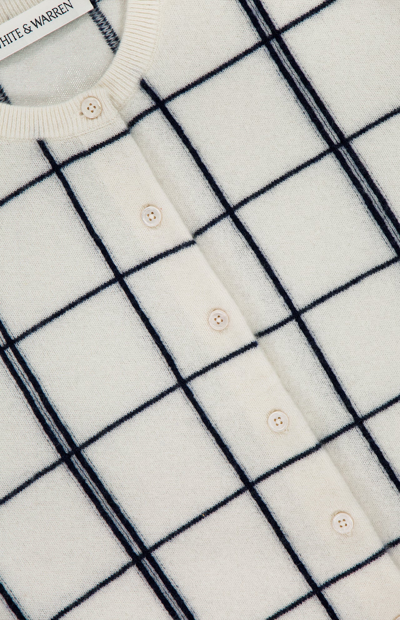 Close-up of the WHITE + WARREN Cashmere Windowpane Cardigan Sweater in off-white, showcasing a black windowpane pattern and front button closure. The collar label displays "WHITE + WARREN.