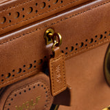 Close-up of the WOLF Brogue Triple Winder with Storage, crafted in brown leather with gold hardware, decorative stitching, and the WOLF brand embossed on a leather tag and circular detail—a sophisticated accessory that perfectly complements your luxury watch winder or stylish watch storage collection.