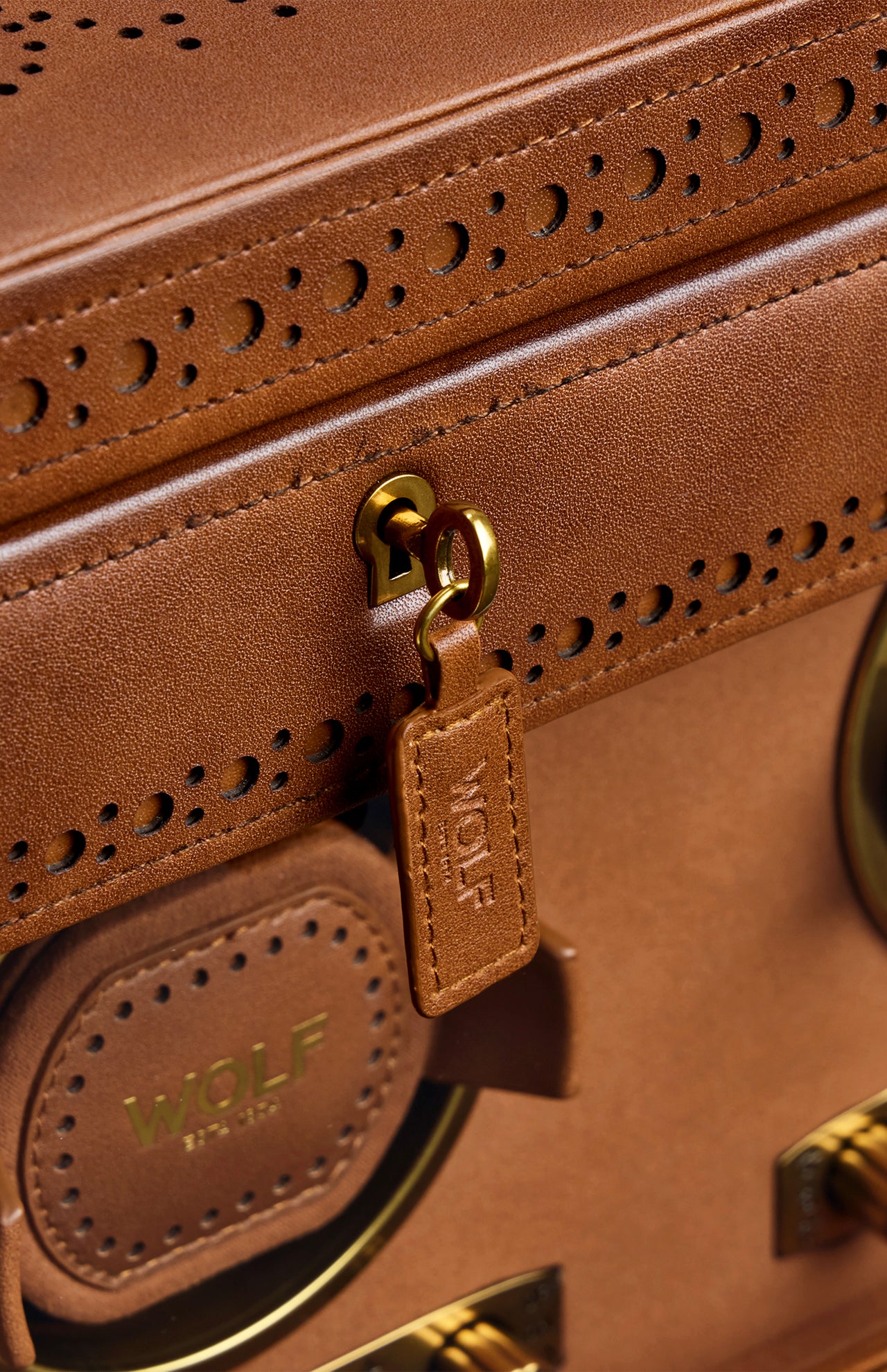 Close-up of the WOLF Brogue Triple Winder with Storage, crafted in brown leather with gold hardware, decorative stitching, and the WOLF brand embossed on a leather tag and circular detail—a sophisticated accessory that perfectly complements your luxury watch winder or stylish watch storage collection.