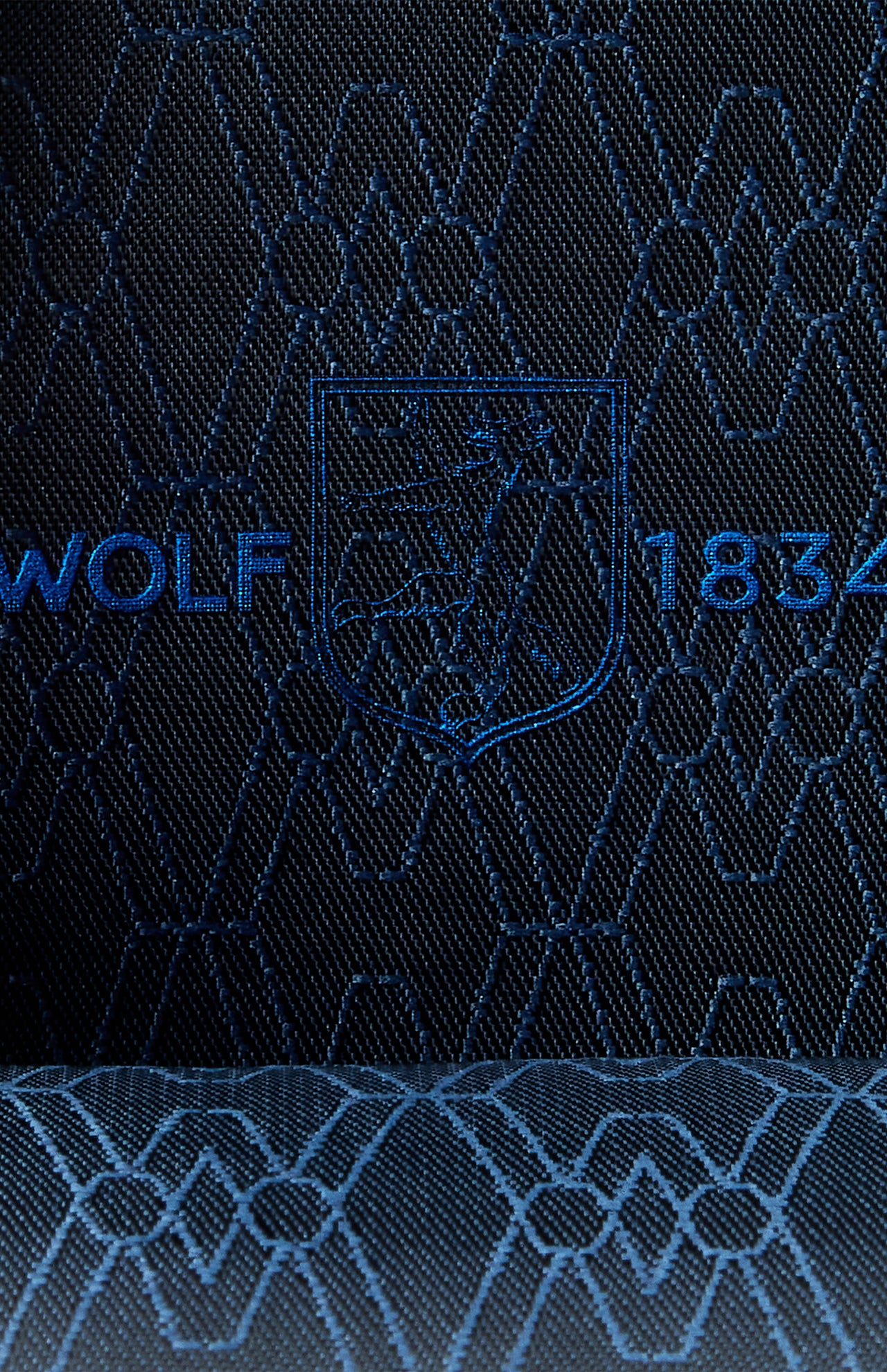 Close-up of dark apple leather fabric with an embroidered crest featuring a wolf, geometric designs, and the text "WOLF 1834" in blue stitching—an elegant detail found on the WOLF Earth Triple Watch Roll, a premium watch storage accessory.