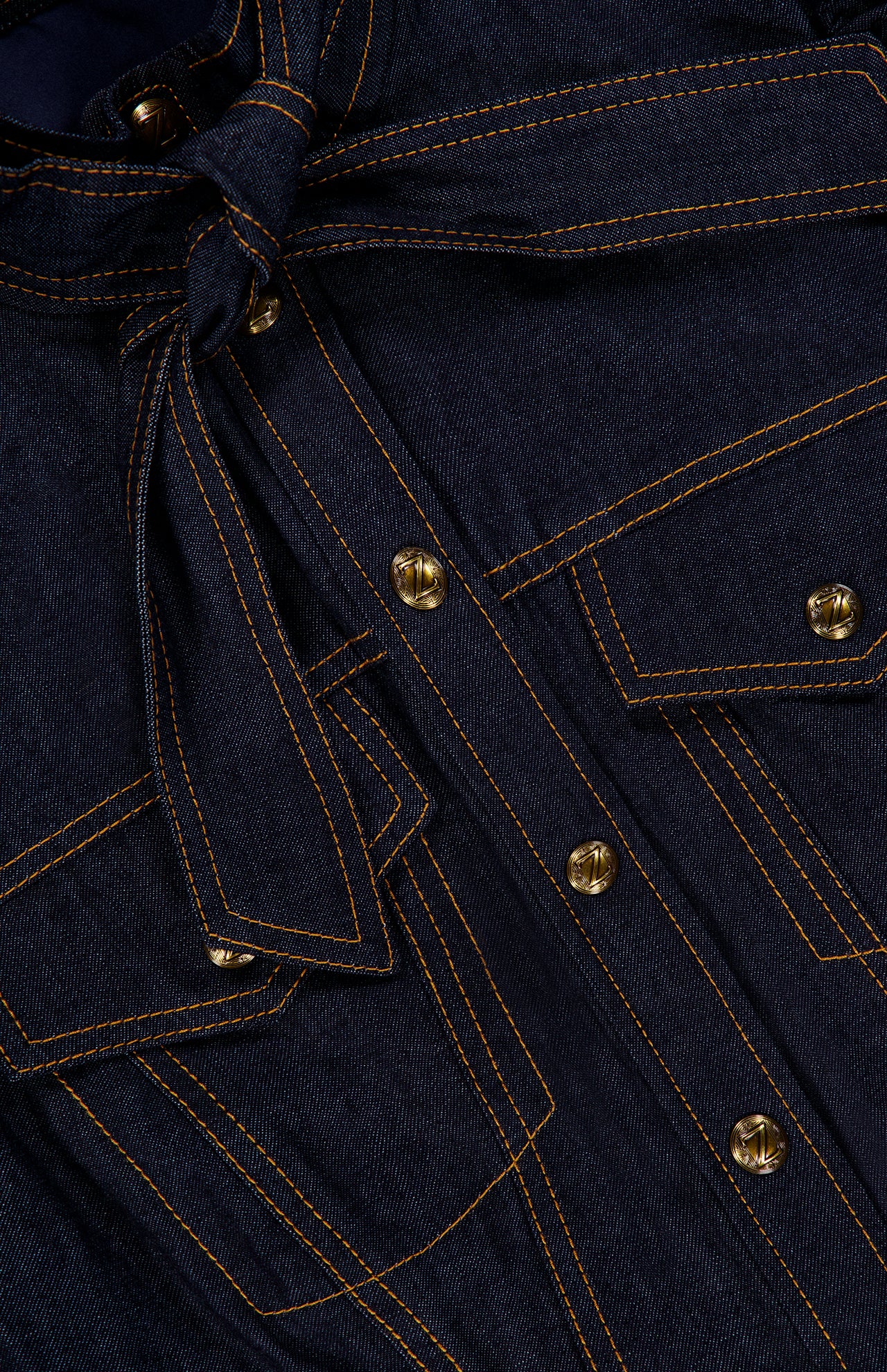 Close-up of deep blue denim fabric featuring orange stitching, metal buttons, flap pockets, and a tied belt at the waist, highlighting the classic corset details of the ZIMMERMANN Denim Corset Mini Dress.