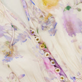 Close-up of the ZIMMERMANN Luna Ruffle Floral Playsuit, featuring semi-sheer fabric with pastel purple, yellow, and green florals. Fabric-covered buttons and drawstring ties at the neckline add a soft, feminine touch.