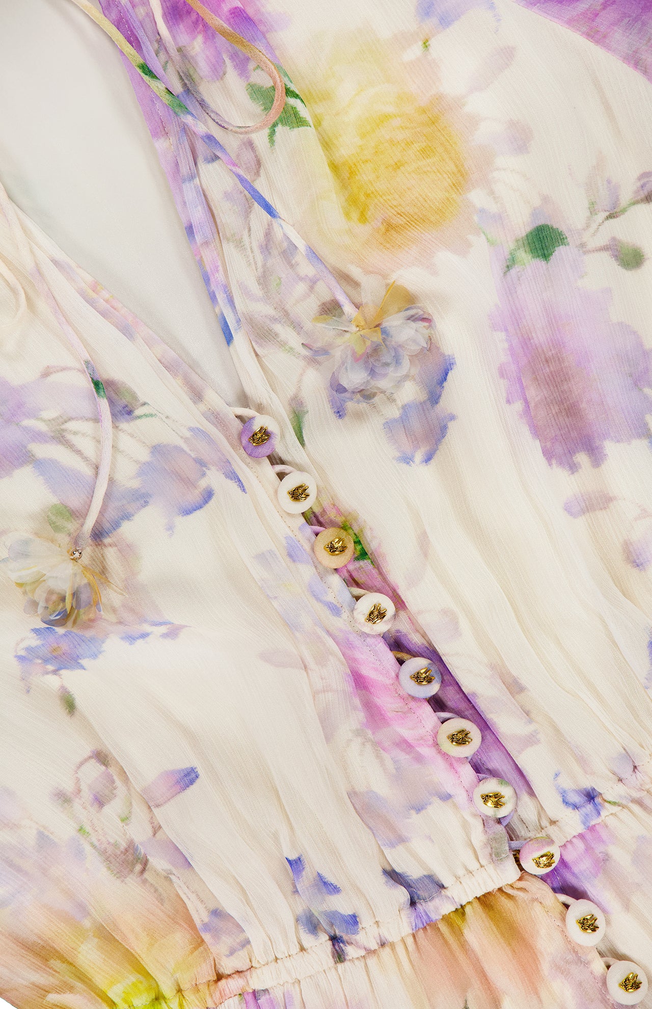 Close-up of the ZIMMERMANN Luna Ruffle Floral Playsuit, featuring semi-sheer fabric with pastel purple, yellow, and green florals. Fabric-covered buttons and drawstring ties at the neckline add a soft, feminine touch.