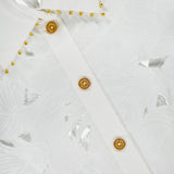Close-up of the Mahon Embroidered Crop Shirt by ZIMMERMANN, featuring ivory cotton with a leaf-like pattern, gold buttons, and gold beading along the collar. The ZIMMERMANN label is visible at the top left.