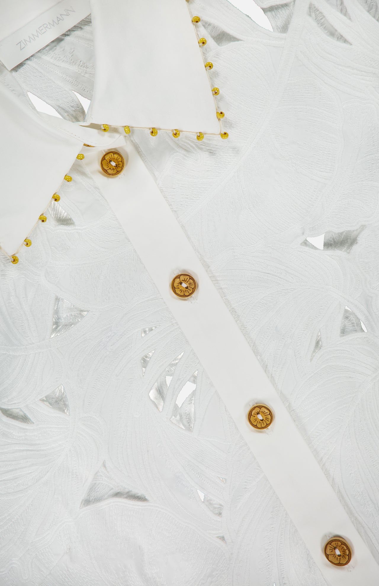 Close-up of the Mahon Embroidered Crop Shirt by ZIMMERMANN, featuring ivory cotton with a leaf-like pattern, gold buttons, and gold beading along the collar. The ZIMMERMANN label is visible at the top left.
