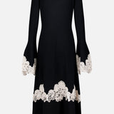 The ZIMMERMANN Valiant Merino Lace Dress is a long black merino wool midi dress with flared sleeves and a gracefully flared skirt, featuring intricate white lace embroidered trim at the cuffs, hem, and near the bottom. It is displayed against a plain white background.