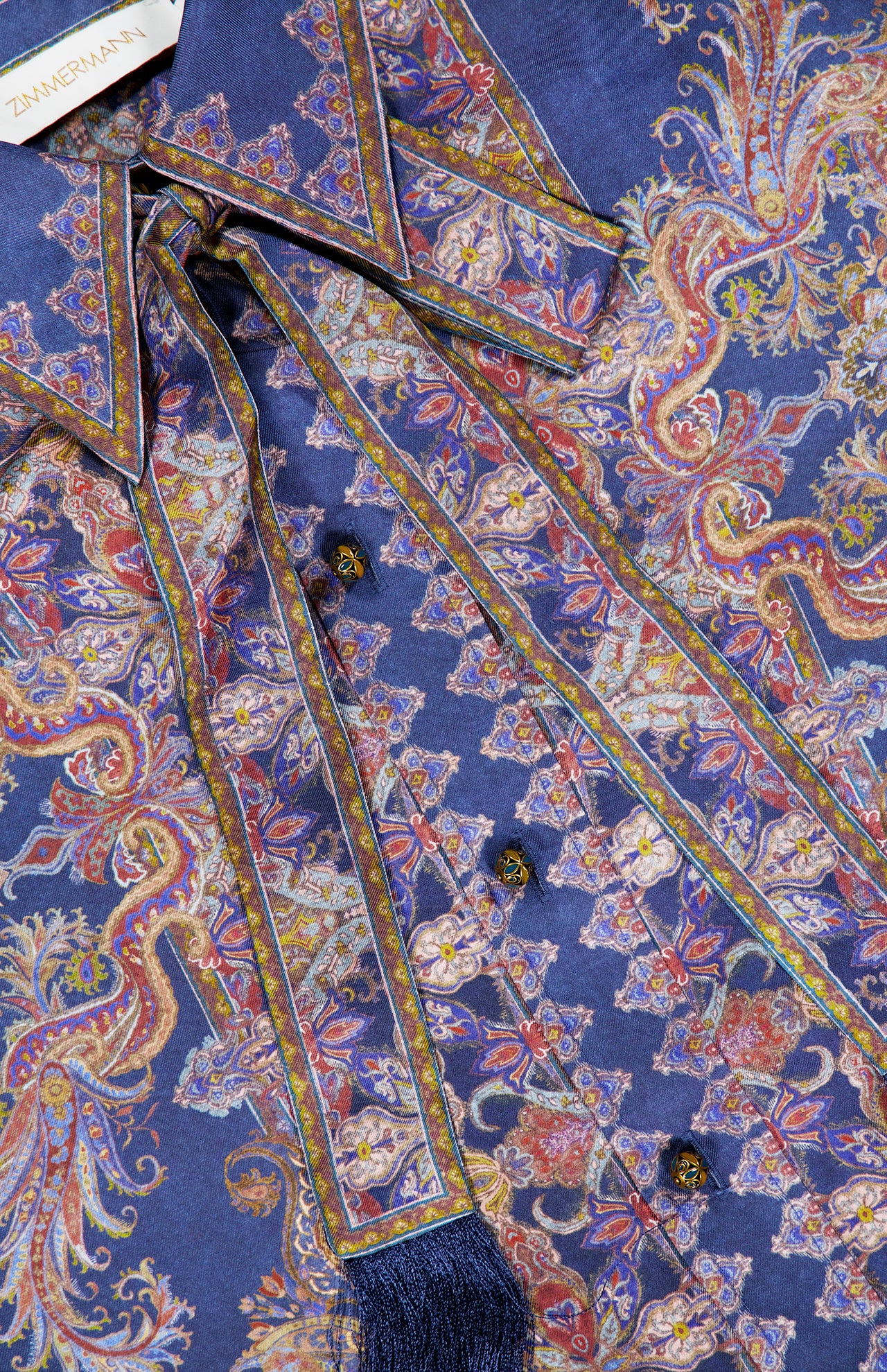 A close-up of the ZIMMERMANN Valiant Relaxed Bow Collar Shirt, featuring a richly patterned blue and gold silk paisley design with ornate buttons, tie details, and the designer’s label on the collar—capturing the essence of this luxurious women’s shirt.