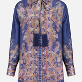The ZIMMERMANN Valiant Relaxed Bow Collar Shirt is a long-sleeve, button-up silk blouse in deep blue, featuring an ornate gold and pink paisley pattern and a matching bow collar necktie embellished with fringed tassels—offering the perfect blend of designer women’s shirt elegance and subtle bohemian charm.