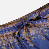 The Valiant Paisley Print Track Pant by ZIMMERMANN showcases a navy base embellished with an ornate paisley and floral pattern in gold and purple hues. These high-rise pull-on pants feature an elastic waistband and a fabric bow tie at the center, combining comfort with elevated style.