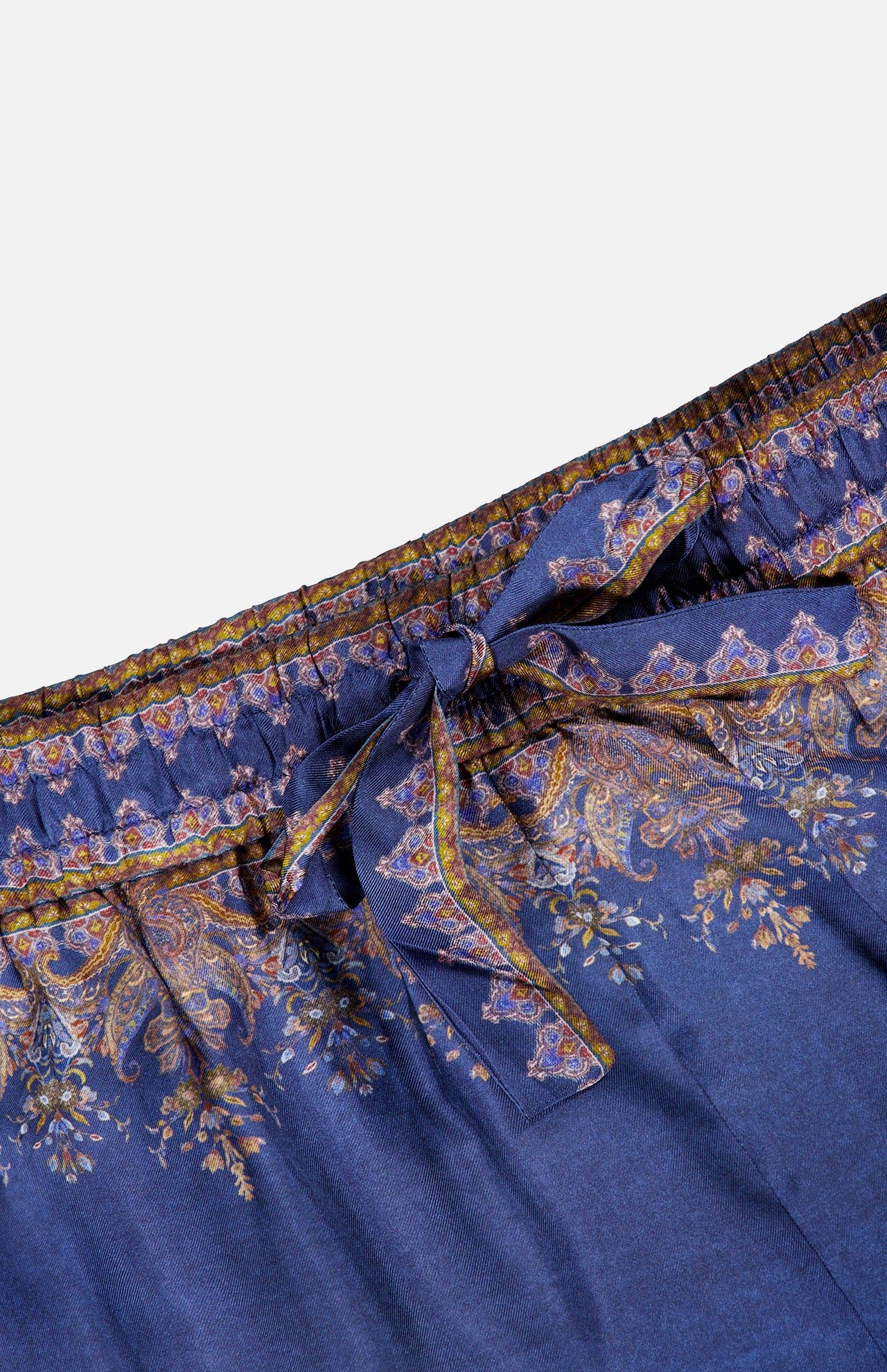 The Valiant Paisley Print Track Pant by ZIMMERMANN showcases a navy base embellished with an ornate paisley and floral pattern in gold and purple hues. These high-rise pull-on pants feature an elastic waistband and a fabric bow tie at the center, combining comfort with elevated style.