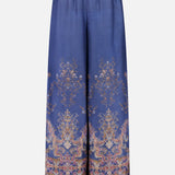 The Valiant Paisley Print Track Pant by ZIMMERMANN features a wide-leg silhouette in silky blue fabric, adorned with an ornate gold and pink paisley floral pattern. These high-rise pull-on pants include an elastic waistband with a drawstring tie, and showcase intricate, symmetrical detailing at both the hem and waist.