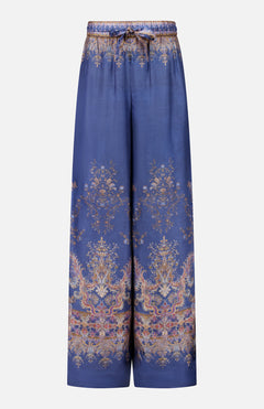 The Valiant Paisley Print Track Pant by ZIMMERMANN features a wide-leg silhouette in silky blue fabric, adorned with an ornate gold and pink paisley floral pattern. These high-rise pull-on pants include an elastic waistband with a drawstring tie, and showcase intricate, symmetrical detailing at both the hem and waist.