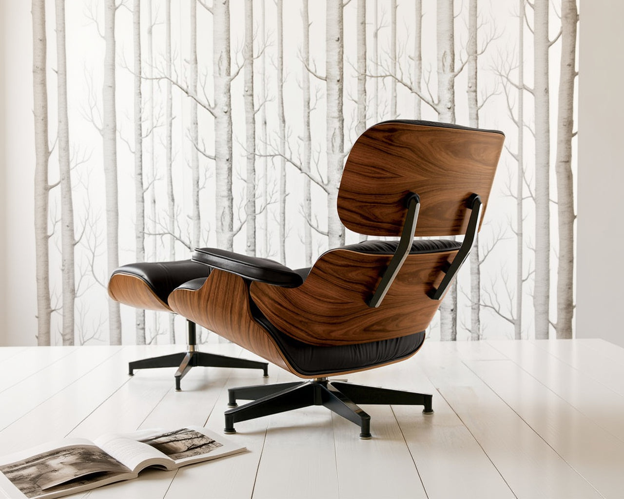 A HERMAN MILLER Eames Lounge Chair and Ottoman with curved Santos Palisander wood and black leather sits on a white floor before birch tree wallpaper; an open book lies nearby.