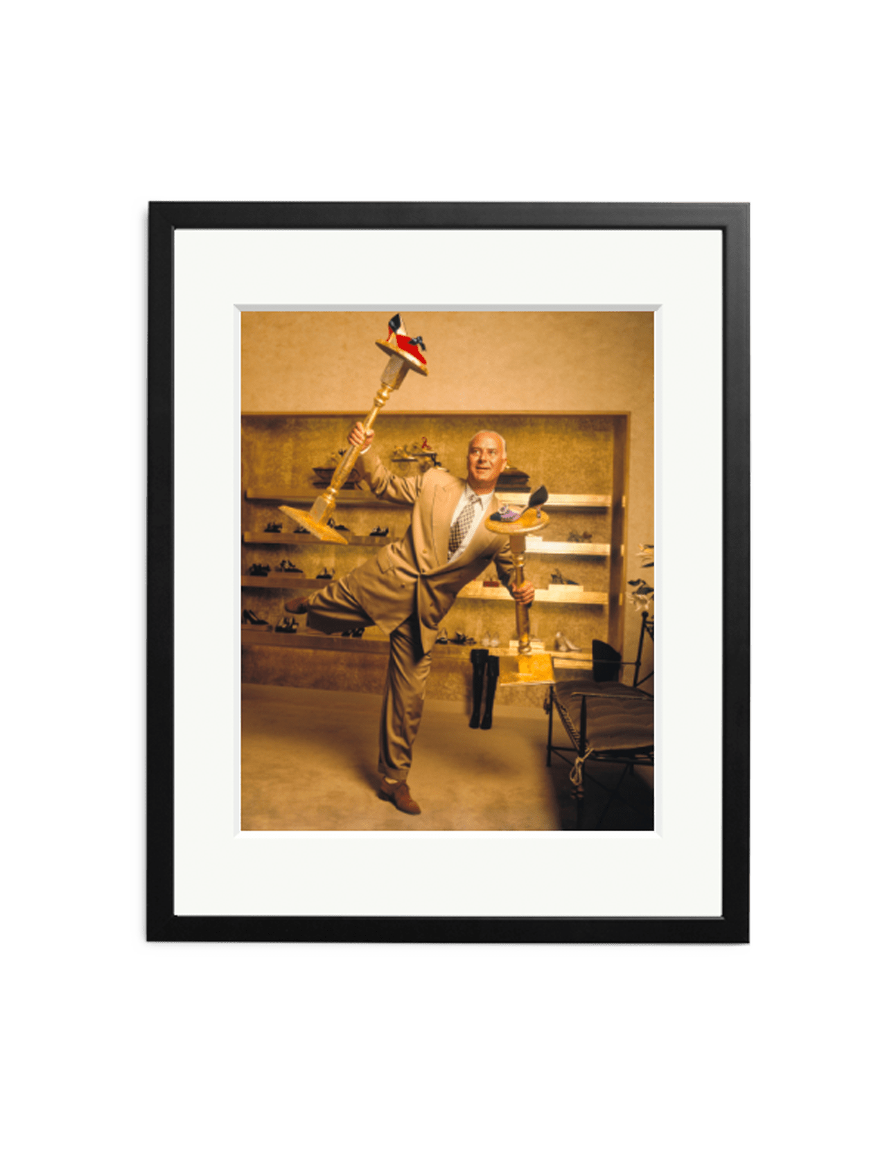 A man in a beige suit balances on one foot in a shoe store, holding several shoes and a Manolo Blahnik gold shoe display. This SONIC EDITIONS photography print features a white mat and black frame.