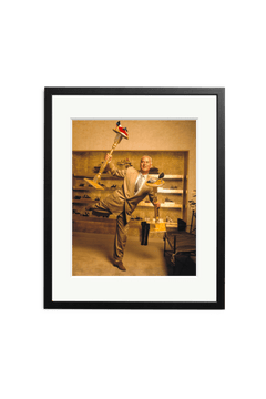 A man in a beige suit balances on one foot in a shoe store, holding several shoes and a Manolo Blahnik gold shoe display. This SONIC EDITIONS photography print features a white mat and black frame.