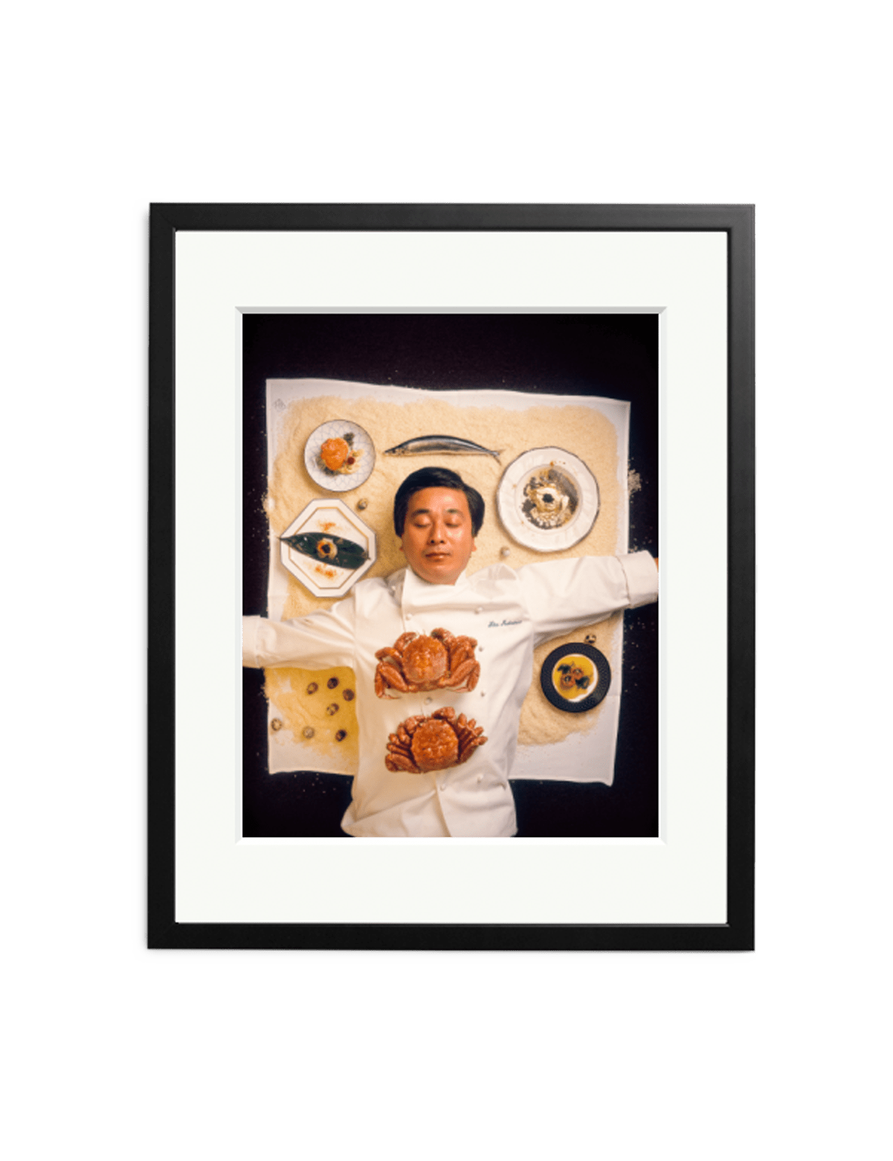 The "Nobu" limited edition by SONIC EDITIONS features a chef in white lying on a table, arms outstretched amid plated dishes and baked bread, artistically arranged. The iconic photo comes framed with a black border and white mat.